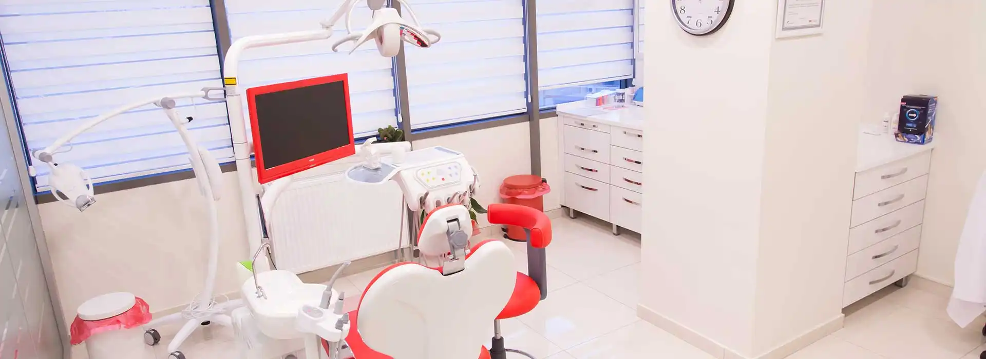 Dent BB kliniğinde bir tedavi odası: kırmızı-beyaz hasta koltuğu, ünit ve gün ışığı alan pencereler
