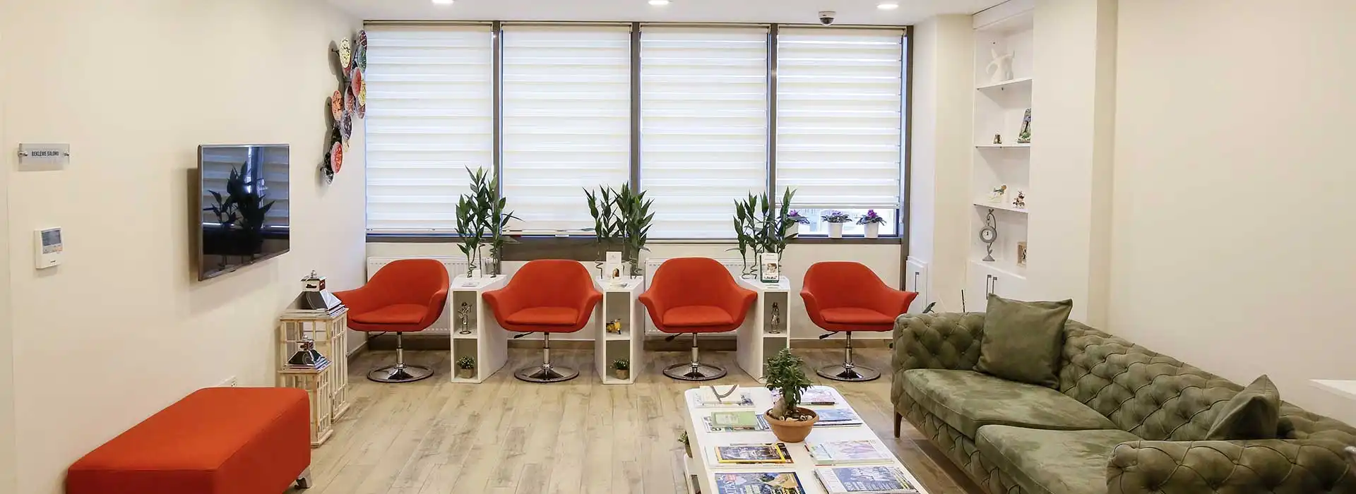 Dent BB kliniğinin bekleme salonuna geniş bir bakış: gün ışığı alan pencereler, kırmızı koltuklar ve yeşil bir kanepe